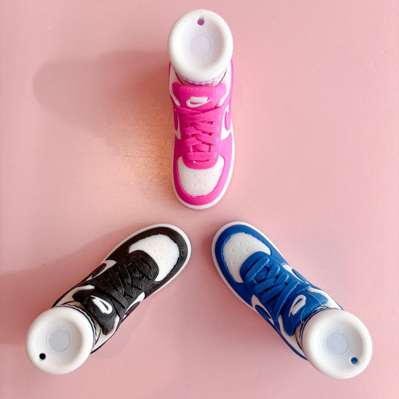 Three pairs of 3d printed sneakers in pink, blue, and black on a pink platform with a checkered wall background.