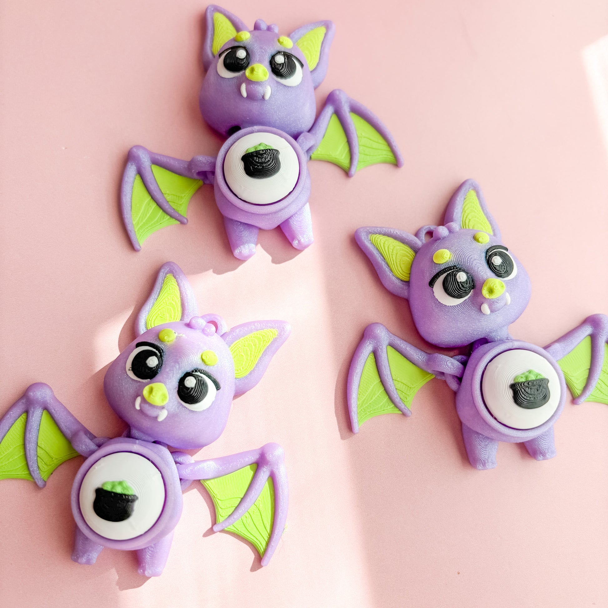 Three purple bat-shaped fidget clicker figurines with green wings on a pink background