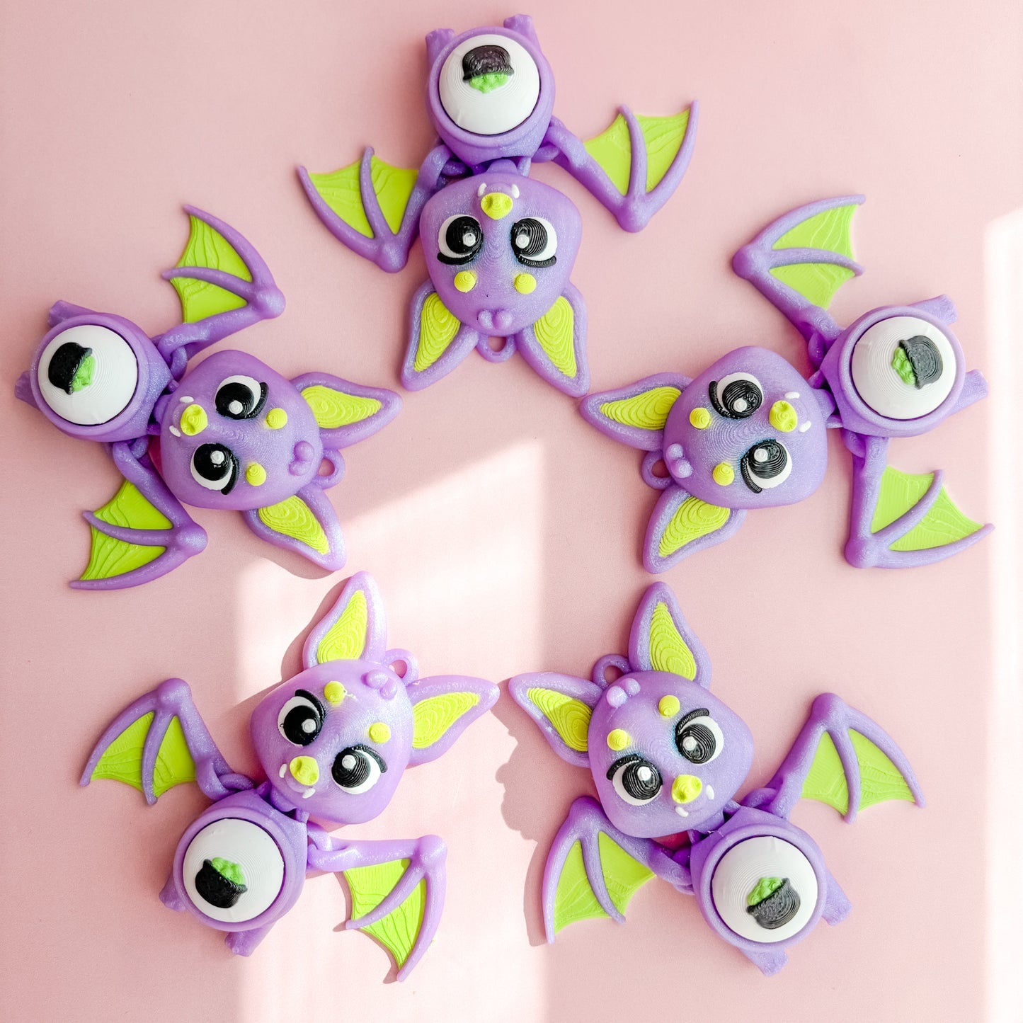 Three purple bat-shaped fidget clicker figurines with green wings on a pink background