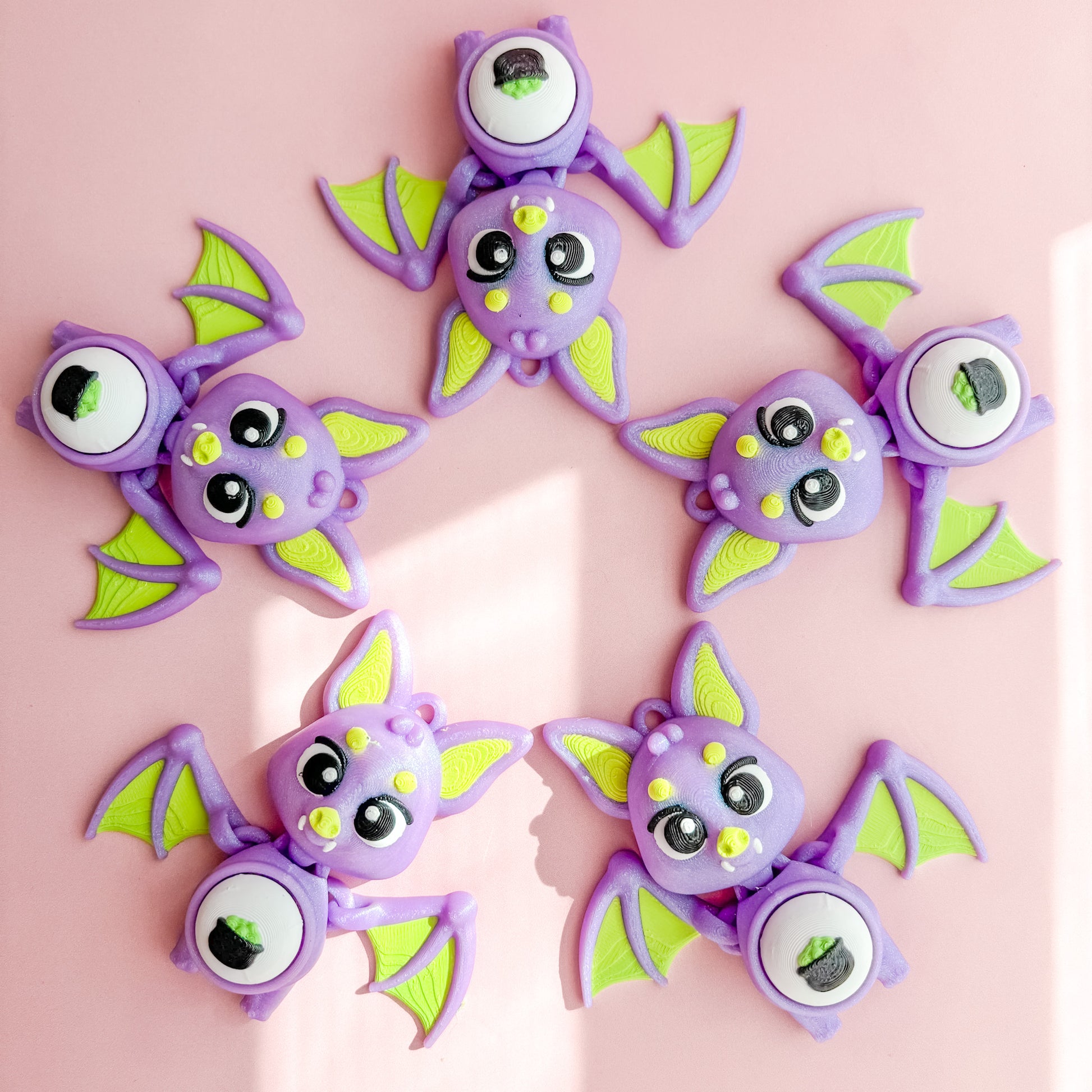 Three purple bat-shaped fidget clicker figurines with green wings on a pink background