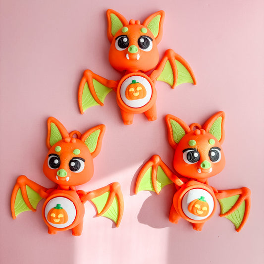 Three orange bat-shaped fidget clicker figurines with green wings on a pink background