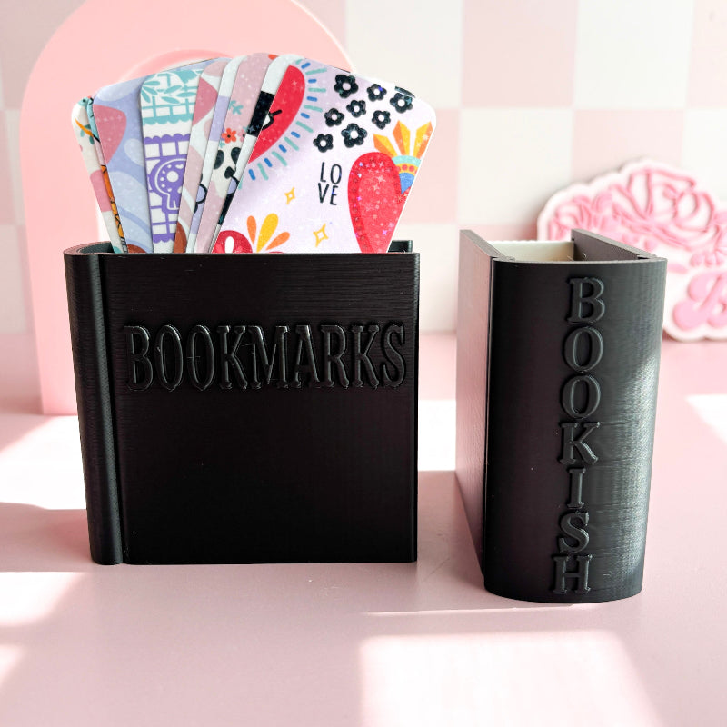 Black 'BOOKMARKS' holder with colorful bookmarks and a black 'BOOKISH' notebook on a pink background.