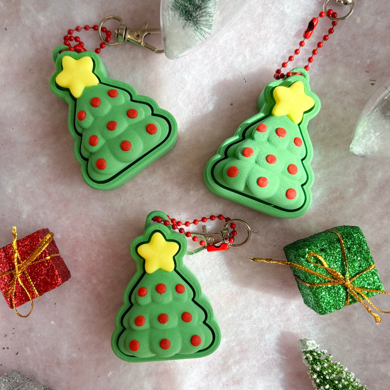 Green Christmas tree-shaped fidget clicker ornaments with red dots and yellow stars on a decorative background.