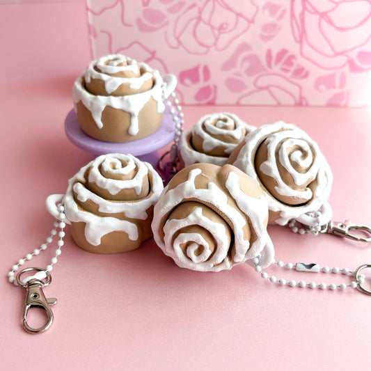 Decorative keychains shaped like cinnamon rolls with white icing on a pink background