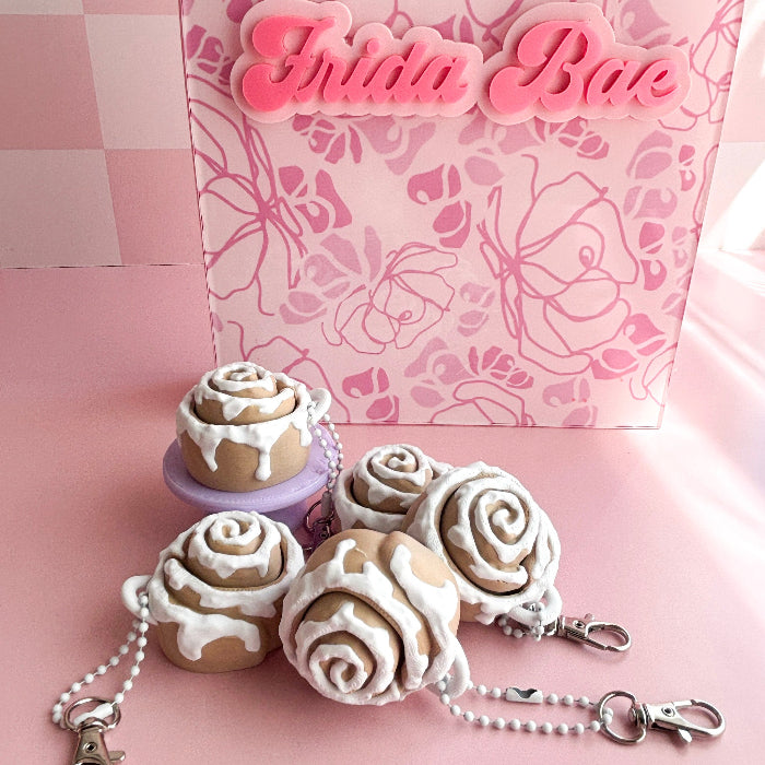 Decorative keychain with cinnamon roll shaped charms in front of a pink box with 'Frida Bae' branding.