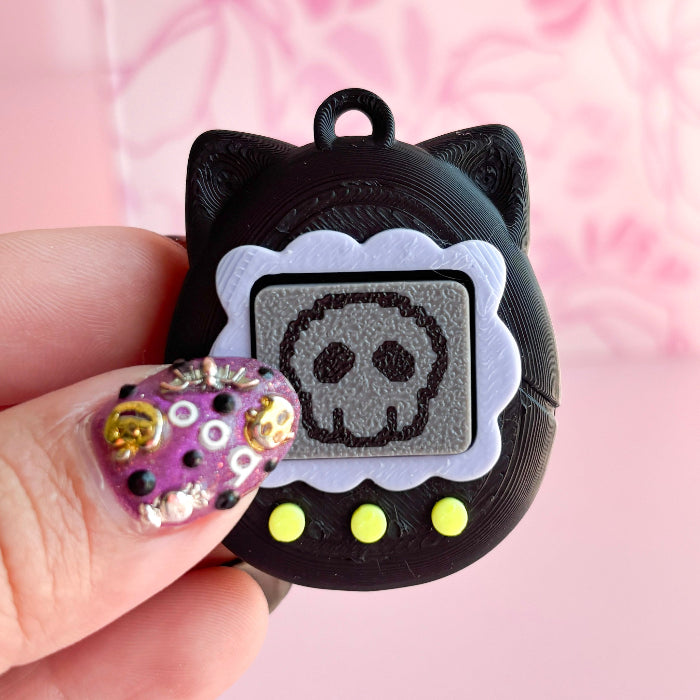 Black cat-shaped charm with skull design held by a hand against a pink background