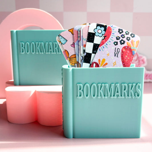 Teal book-shaped container with colorful bookmarks on a pink background