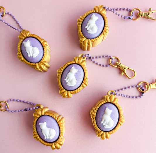 Cat-themed keychains with gold frames and purple backgrounds on a pink background