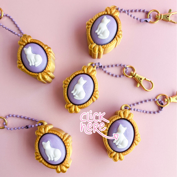 Cat-themed keychains with gold frames and purple backgrounds on a pink background
