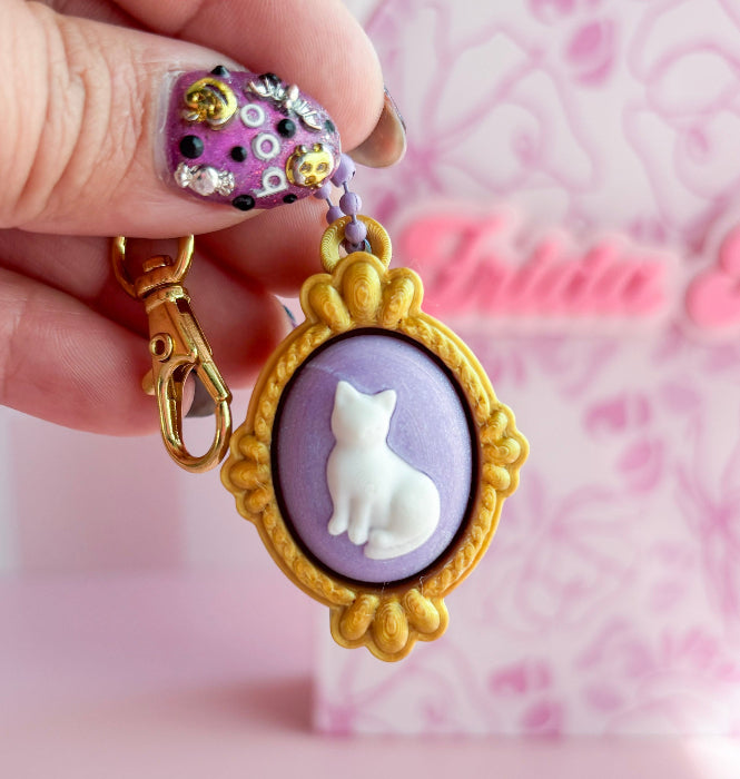 Cat-themed keychain with gold frame and purple background held by a hand against a pink background.