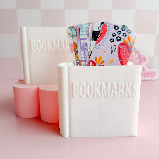 White container with 'BOOKMARKS' text holding colorful bookmarks on a pink background