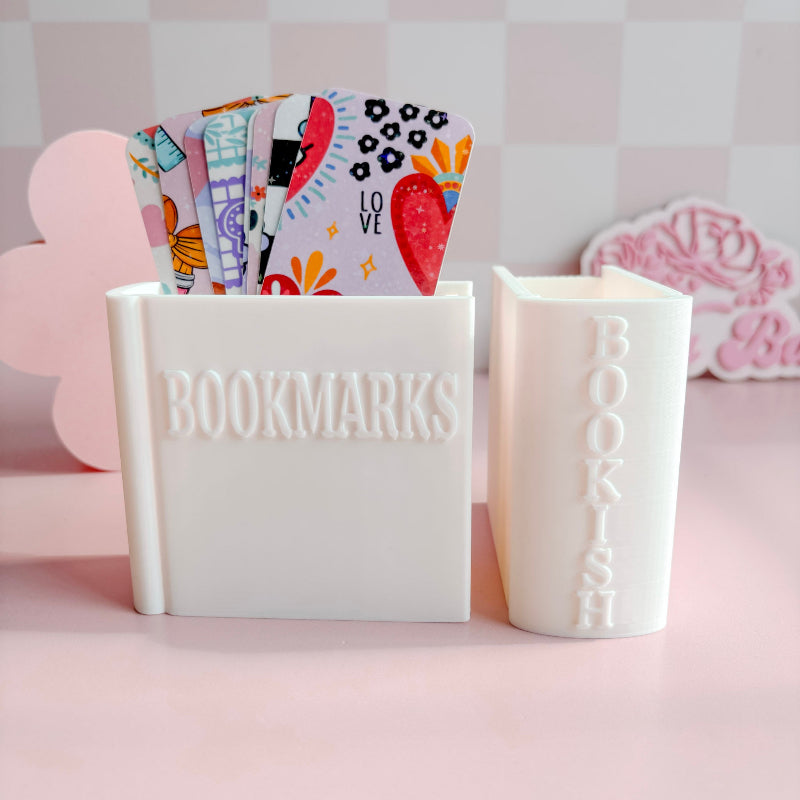 White container with 'BOOKMARKS' and a smaller container with 'BOOKISH', holding colorful bookmarks on a pink background.