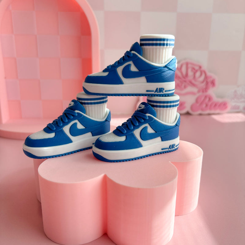Three pairs of 3d printed sneakers in pink, blue, and black on a pink platform with a checkered wall background.