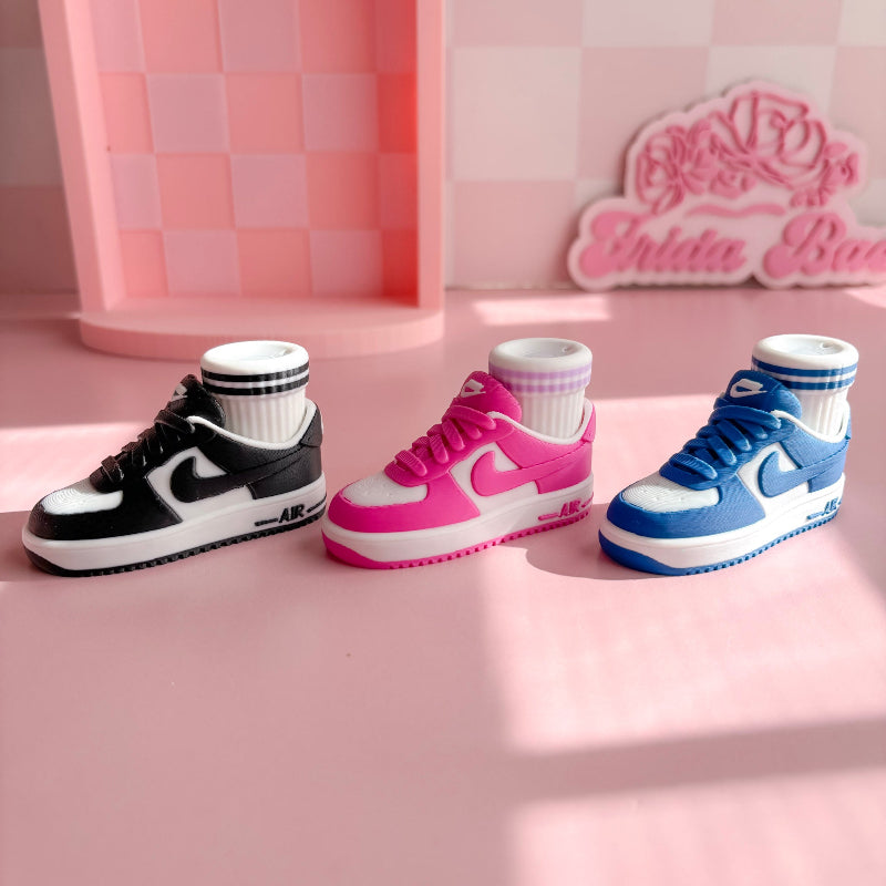 Three pairs of 3d printed sneakers in pink, blue, and black on a pink platform with a checkered wall background.
