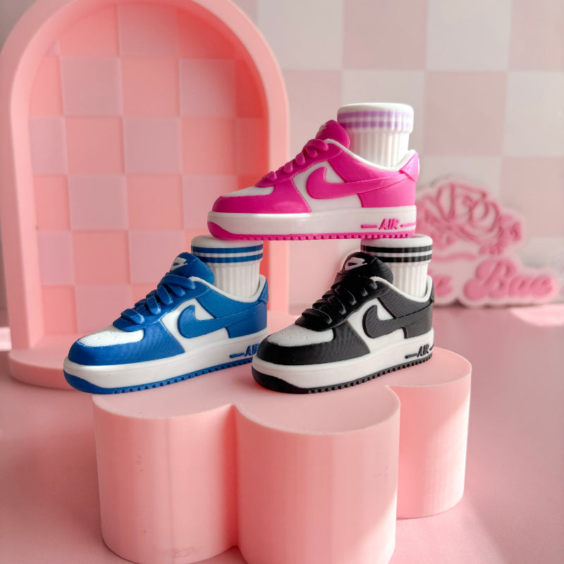Three pairs of 3d printed sneakers in pink, blue, and black on a pink platform with a checkered wall background.