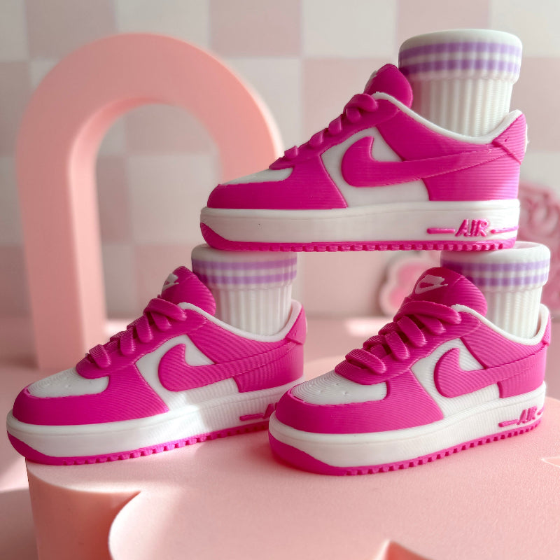 Three pairs of 3d printed sneakers in pink, blue, and black on a pink platform with a checkered wall background.