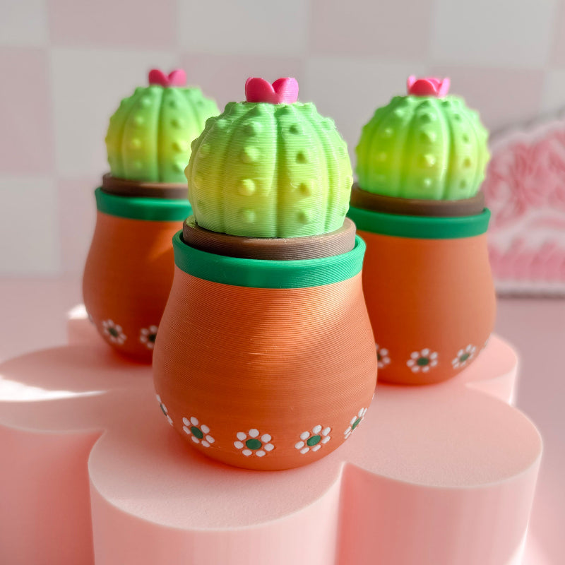 Three cactus-shaped candles in terracotta pots on a pink surface with a blurred background.