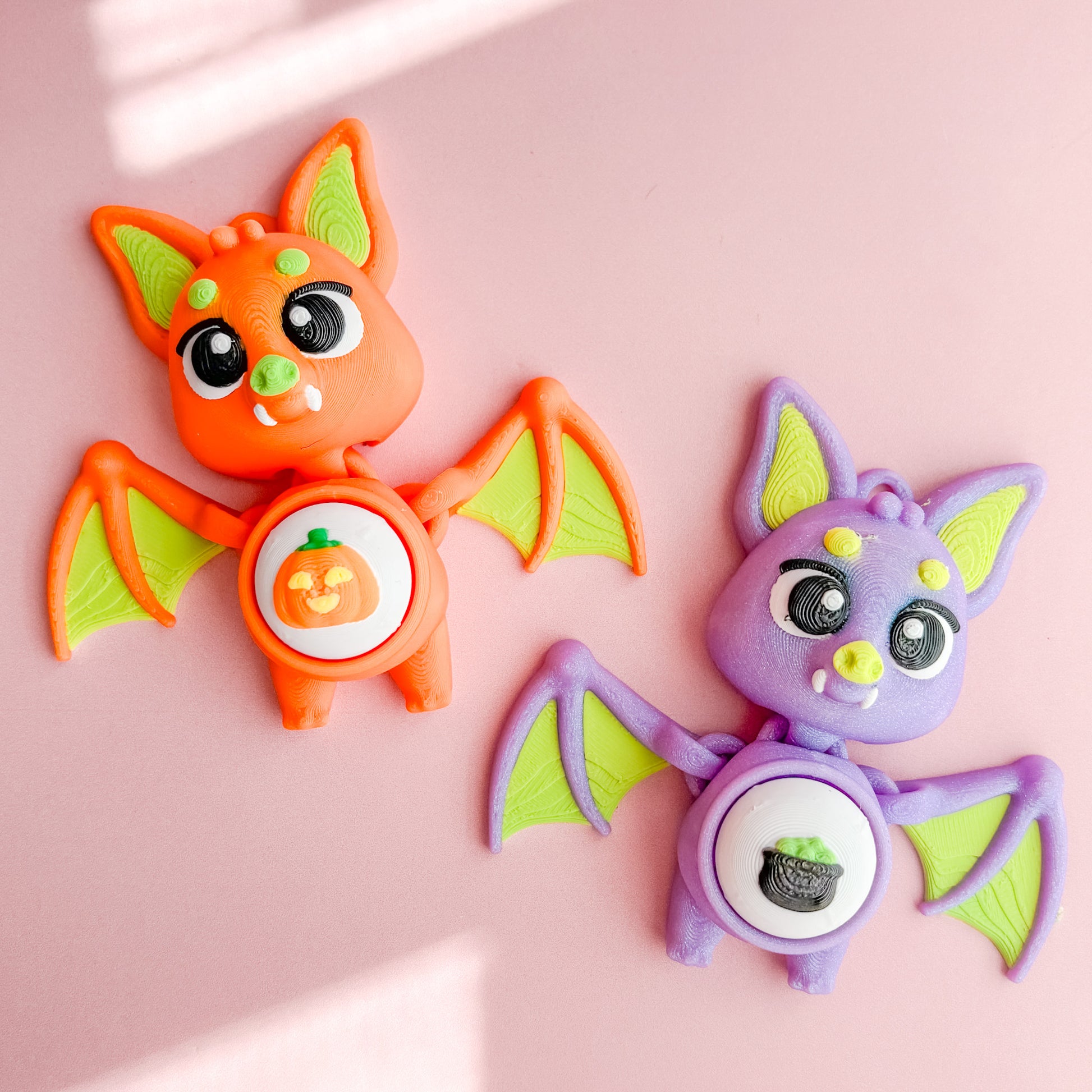 Two bat-shaped fidget clicker figurines with green wings on a pink background