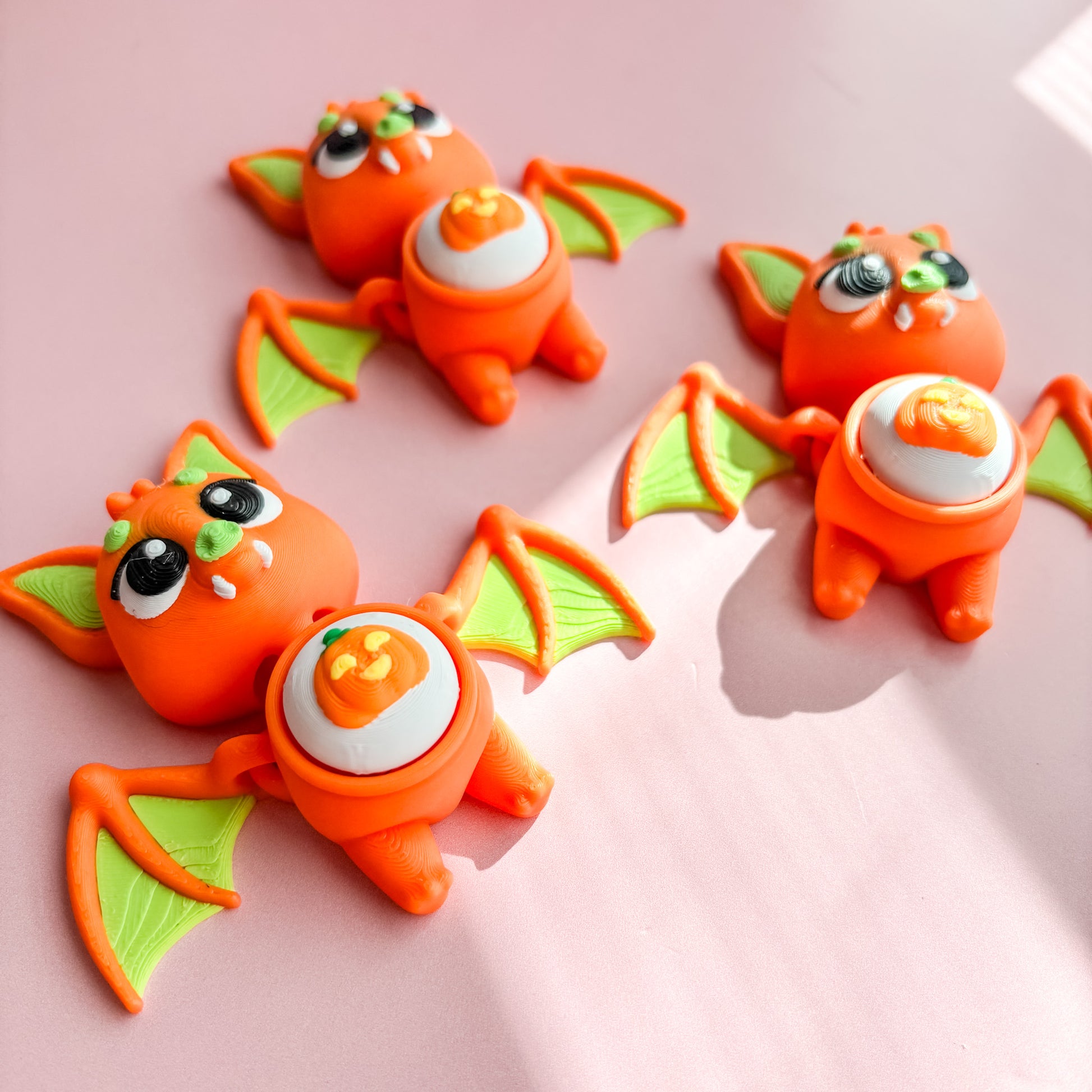Three orange bat-shaped fidget clicker figurines with green wings on a pink background