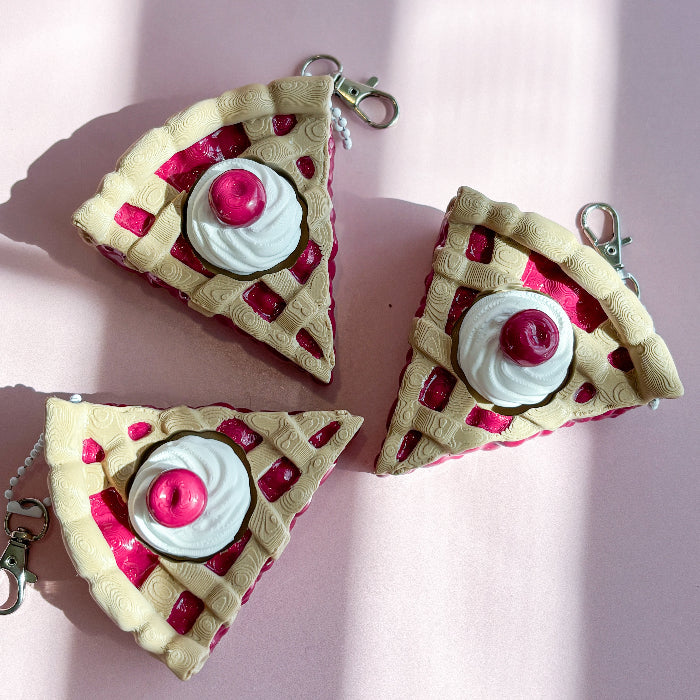 Three cherry pie shaped keychains with pink and red toppings on a pink background