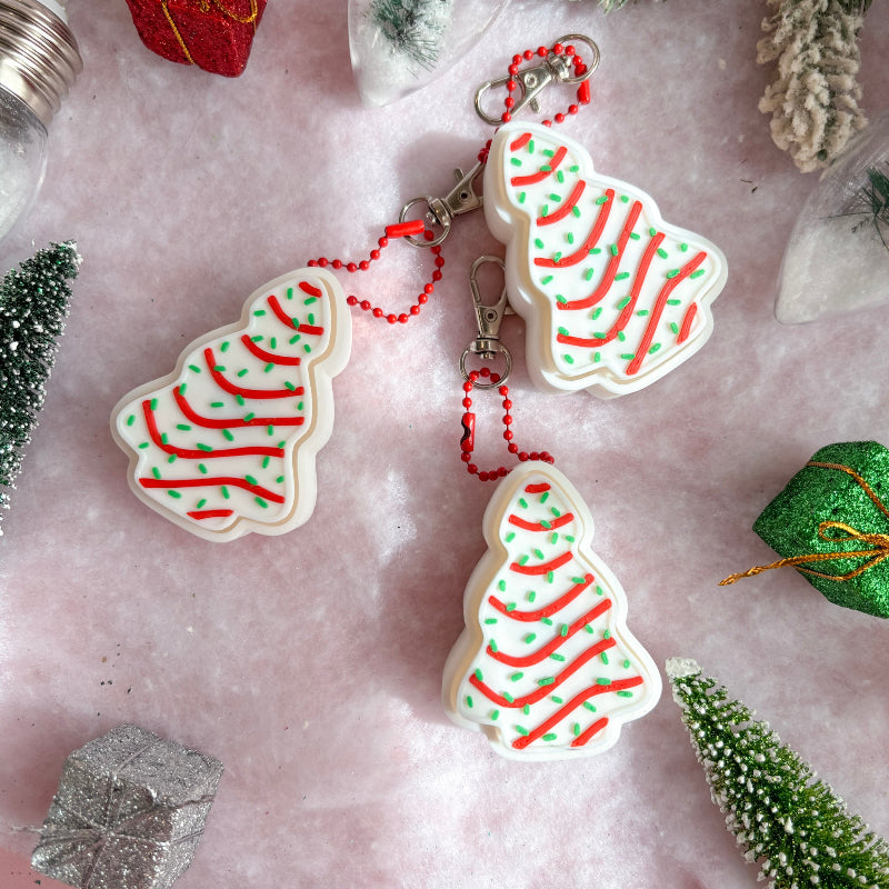 Christmas tree-shaped keychains with red and green patterns on a pink surface with decorative elements.