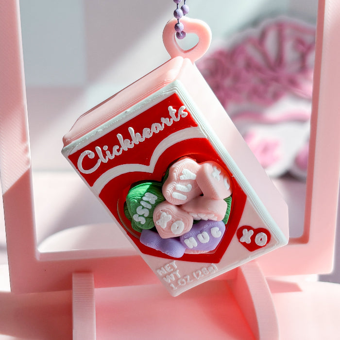Pink and red 'Clickhearts' candy container charm with decorative elements on a pink background.