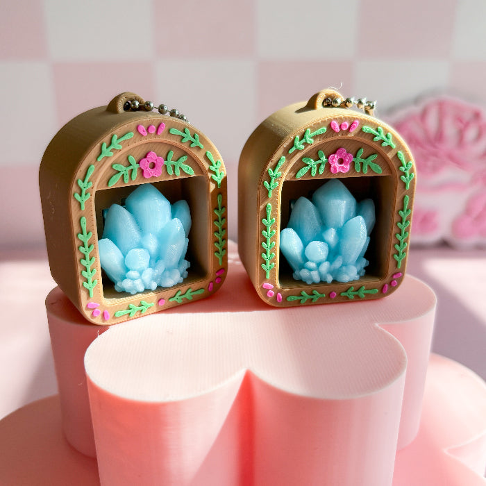 Two decorative keychain fidget clicker with blue crystal-like objects inside wooden frames on a pink surface.