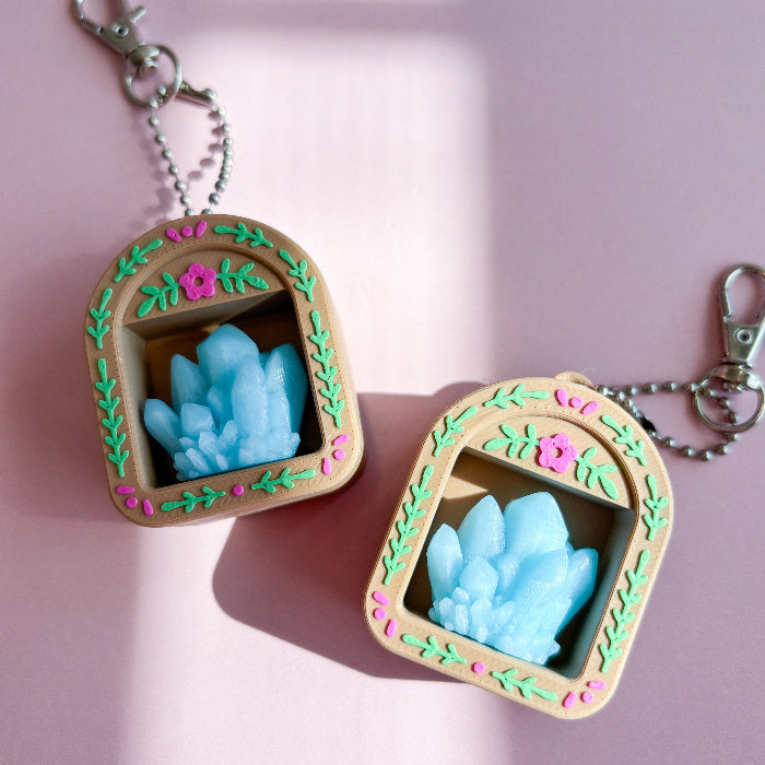 Two decorative keychains with blue crystal-like designs on a pink background