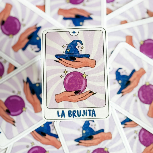 Card from a tarot deck titled 'La Brujita' featuring hands holding a purple sphere and a blue witch's hat.