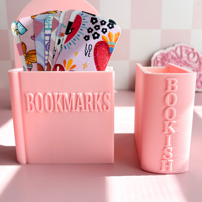Pink container with 'BOOKMARKS' and 'BOOKISH' text, holding colorful bookmarks on a light pink background.