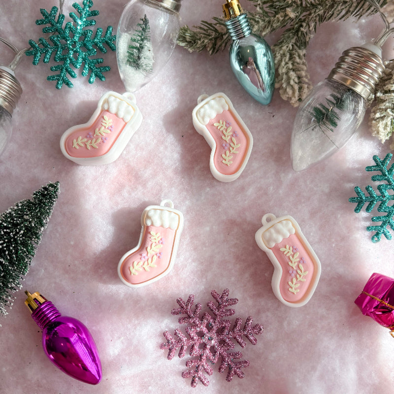 Decorative Christmas ornaments including pink stockings, purple light bulbs, and green snowflakes on a pink surface.