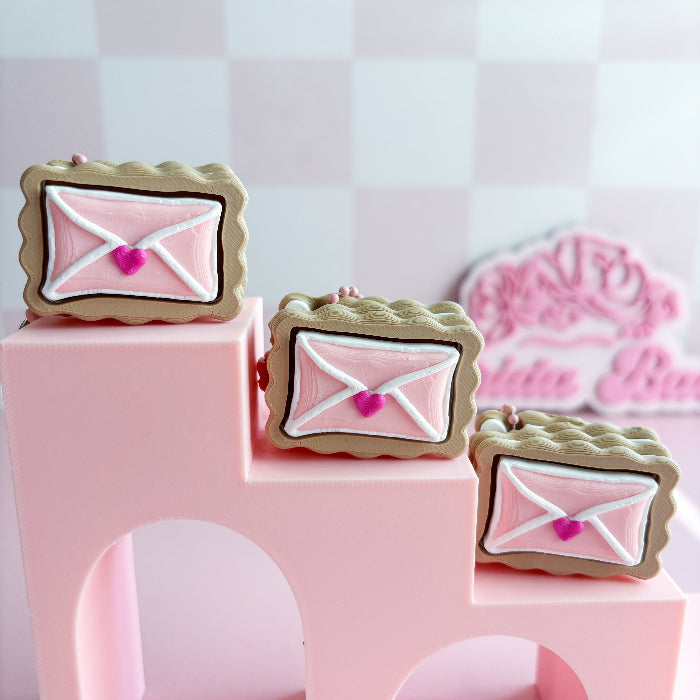 Decorative cookies shaped fidget clicker like pink envelopes with heart details on a pink stand.