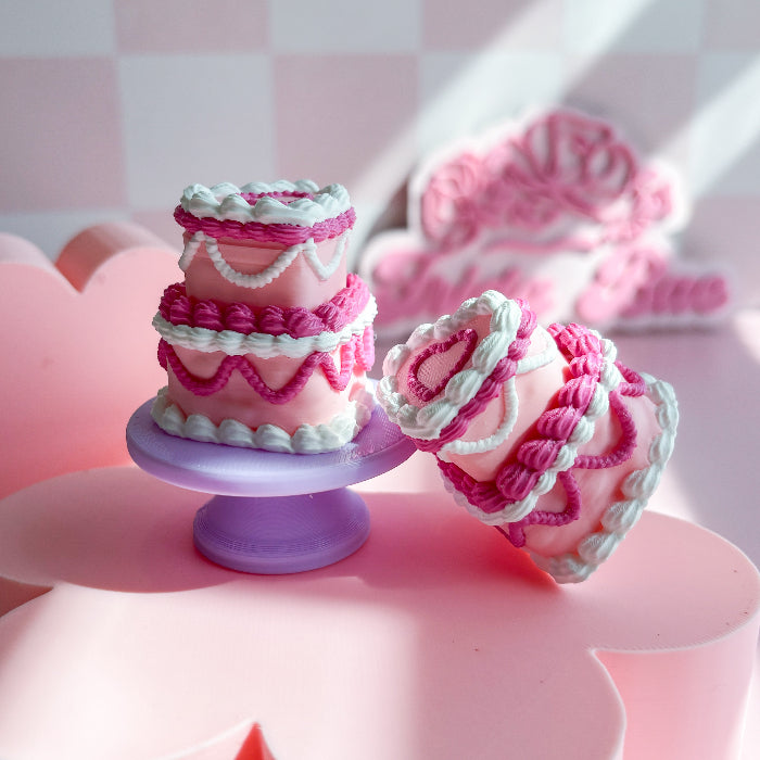 Small pink cake fidget clicker with white frosting on a purple stand against a pink checkered background