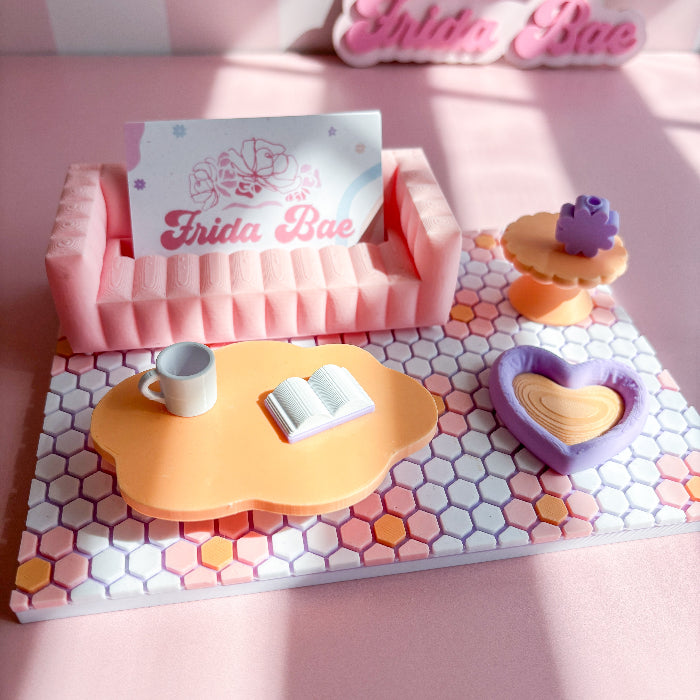 Miniature pink sofa with a book, cup, and heart-shaped object on a colorful surface with 'Frida Bae' branding.