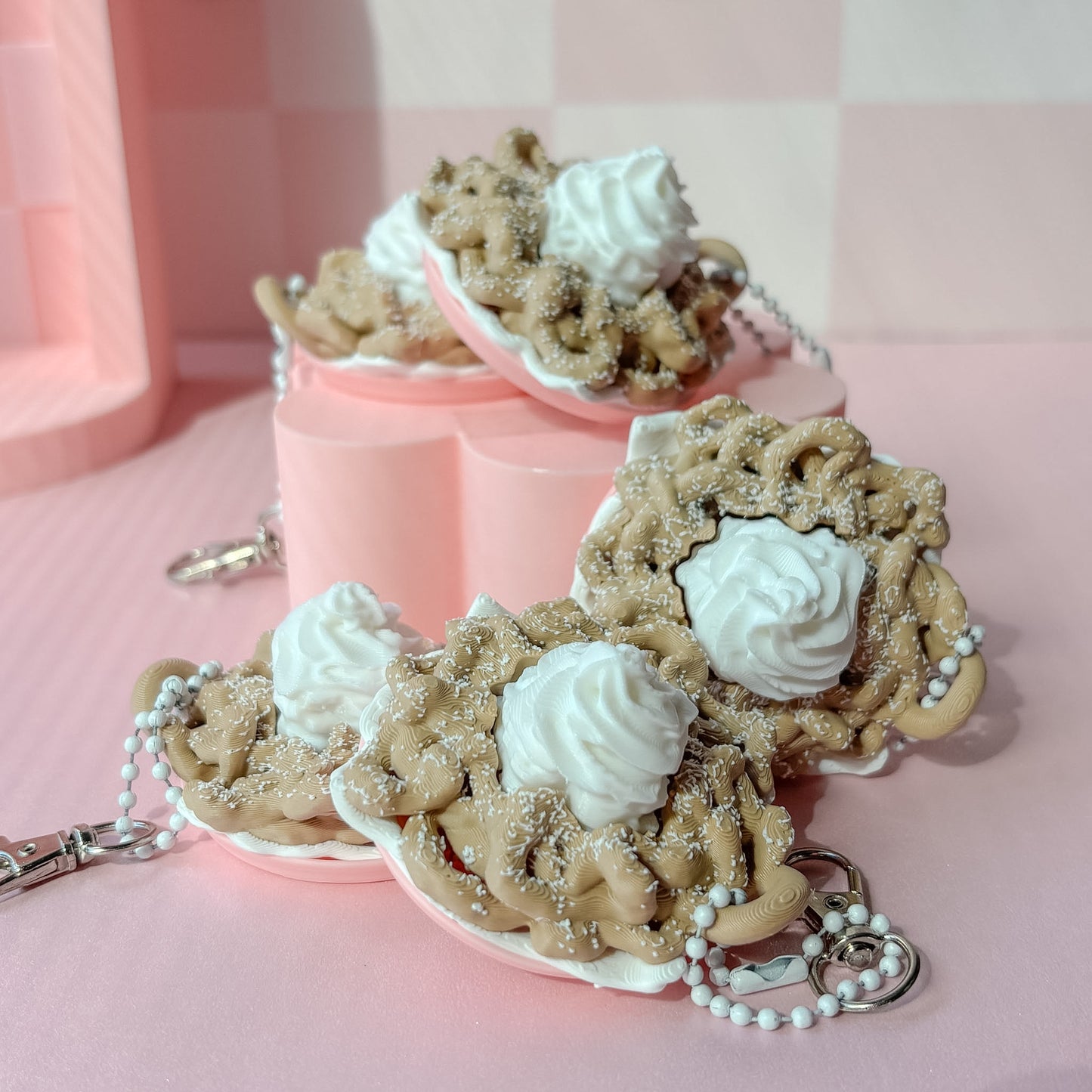 Keychains shaped like funnel cake with white frosting on a pink checkered background