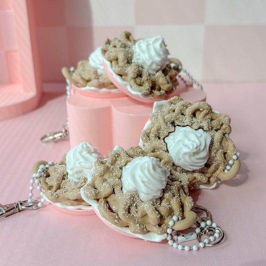 Keychains shaped like funnel cake with white frosting on a pink checkered background