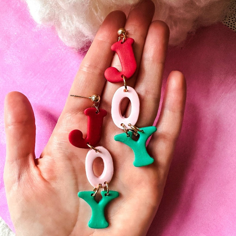 Make some holiday cheer with these JOY Christmas Dangle Earrings! Crafted from durable polymer clay and boasting a merry mix of pink, red, and green, they're sure to become your favorite seasonal accessory. Perfect for a bit of jolly jangling in your ears!